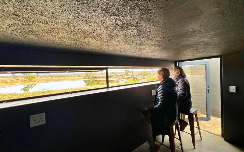 Sian and Vikki of Marula Hill Travel sitting inside the Ximuwu Hide, looking out through a narrow viewing window at the surrounding bush and waterhole.