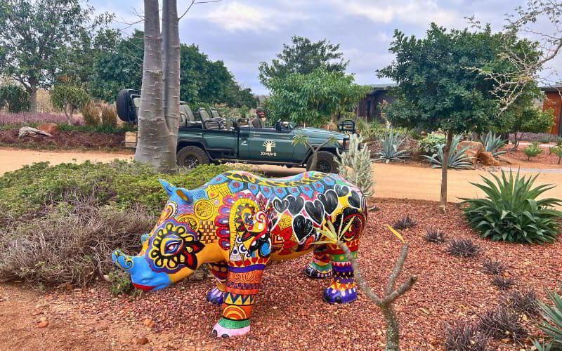 A brightly painted, multicoloured rhino sculpture with intricate patterns and heart motifs stands in a garden at Ximuwu Lodge. In the background, a green Ximuwu game vehicle with a smiling guide is parked on a dirt path surrounded by lush vegetation and succulents.  