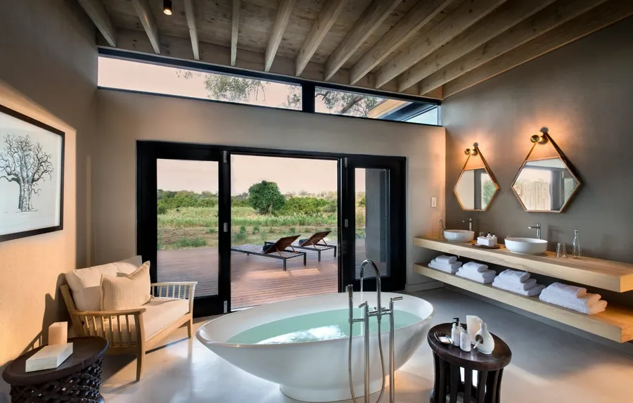 A two-bedroom river suite bathroom at Lion Sands River Lodge — freestanding tub framed by floor-to-ceiling doors open to the Sabie River and bushveld