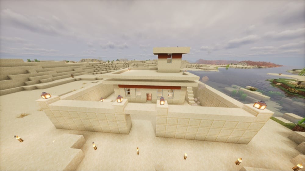 Flat-roofed sandstone house with a red band under the roof and a small rooftop room, enclosed by a low sandstone wall with lantern-topped posts and a gap at the front, with torches in the sand and a lake beside it.