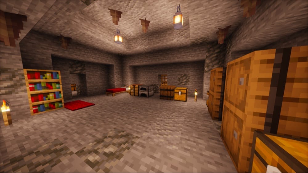 Lit stone cave room furnished with a red bed and carpet, a bookshelf, a crafting table, a furnace, a barrel and chests, with lanterns hanging from the ceiling, torches on the floor, and pointed dripstone overhead.