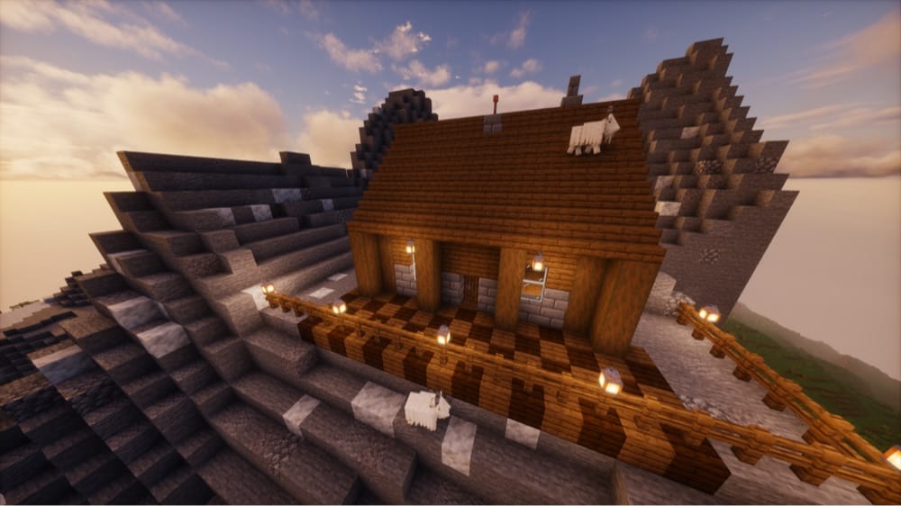 Spruce chalet on a mountain ledge with stone-brick walls, log posts, a checkered plank porch, a fence railing with lanterns, and a lightning rod on the roof, with two goats nearby.
