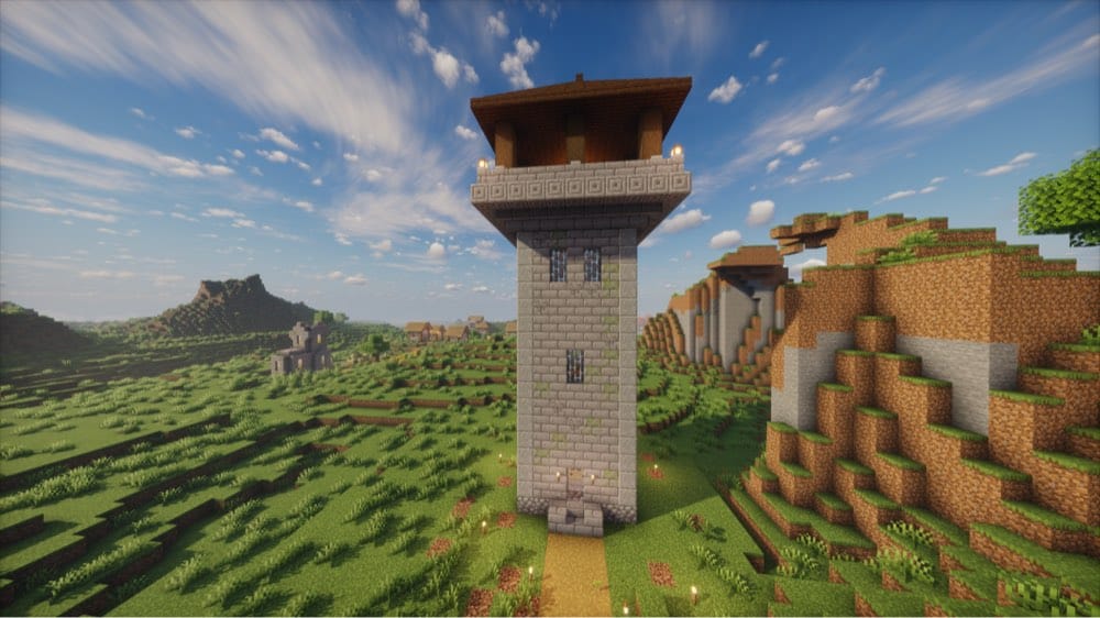 Tall mossy stone-brick watchtower with iron-bar windows and a door at its base, topped by a wider chiseled stone-brick ledge and a wooden-roofed lookout with lanterns, standing on grassland with a village and a hillside behind it.