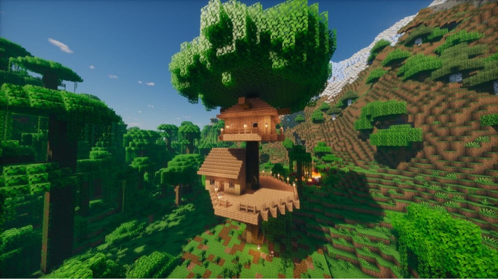 Two-level wooden treehouse built around a thick trunk under a leafy canopy, with a gabled cabin on a wide railed deck below and a smaller cabin with a balcony above, on a forested hillside.