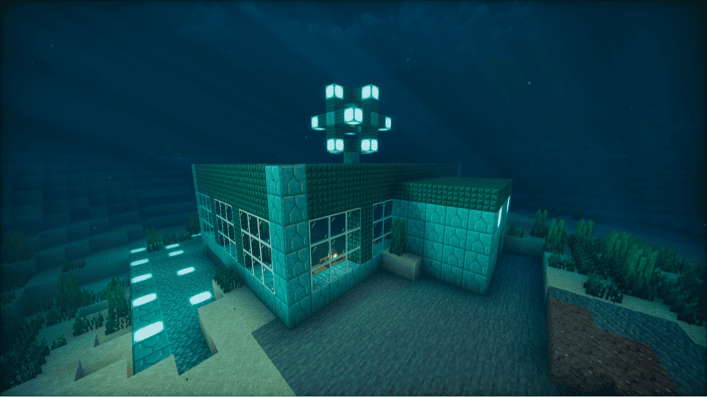 Underwater base on the sea floor with prismarine-brick walls, large glass windows and a dark prismarine roof, a glowing cluster of sea lanterns on top, a sea lantern path on the left, and a lit room visible through the glass.