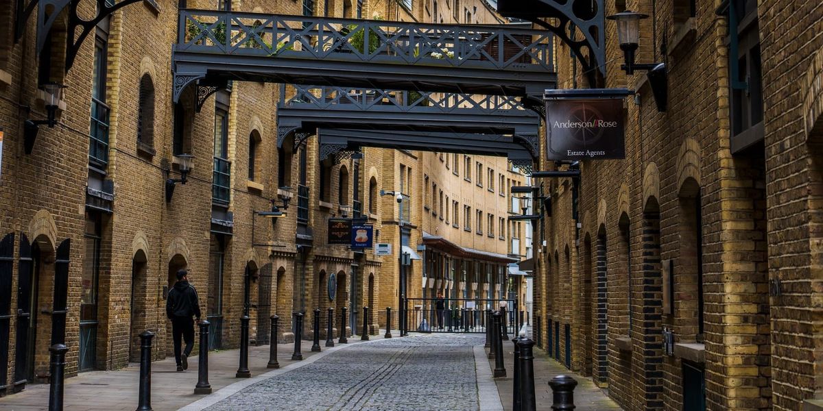 A cobbled Victorian warehouse street in London, with iron overhead walkways linking the brick buildings and a single figure walking between them.