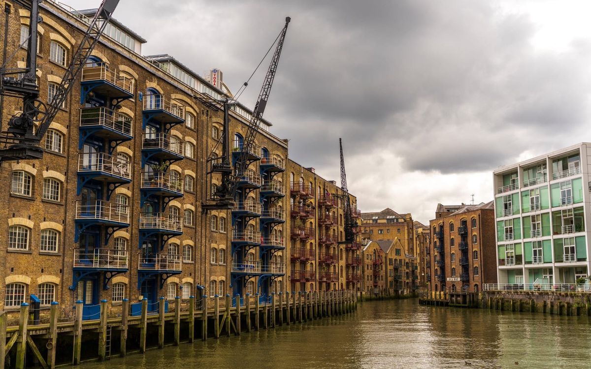 Converted Victorian warehouses along a London river, with old dockside cranes, blue ironwork balconies, wooden pilings in the water, and a modern glass building beside them under a grey sky.