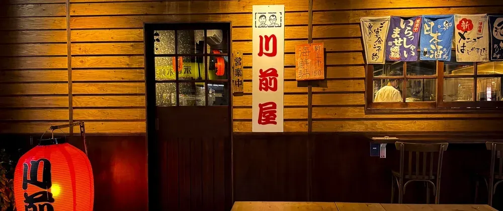 A Japanese izakaya at night: a warmly lit wooden facade with a glowing red paper lantern, a wooden door, vertical signage and hanging banners, and a chef visible through a side window.