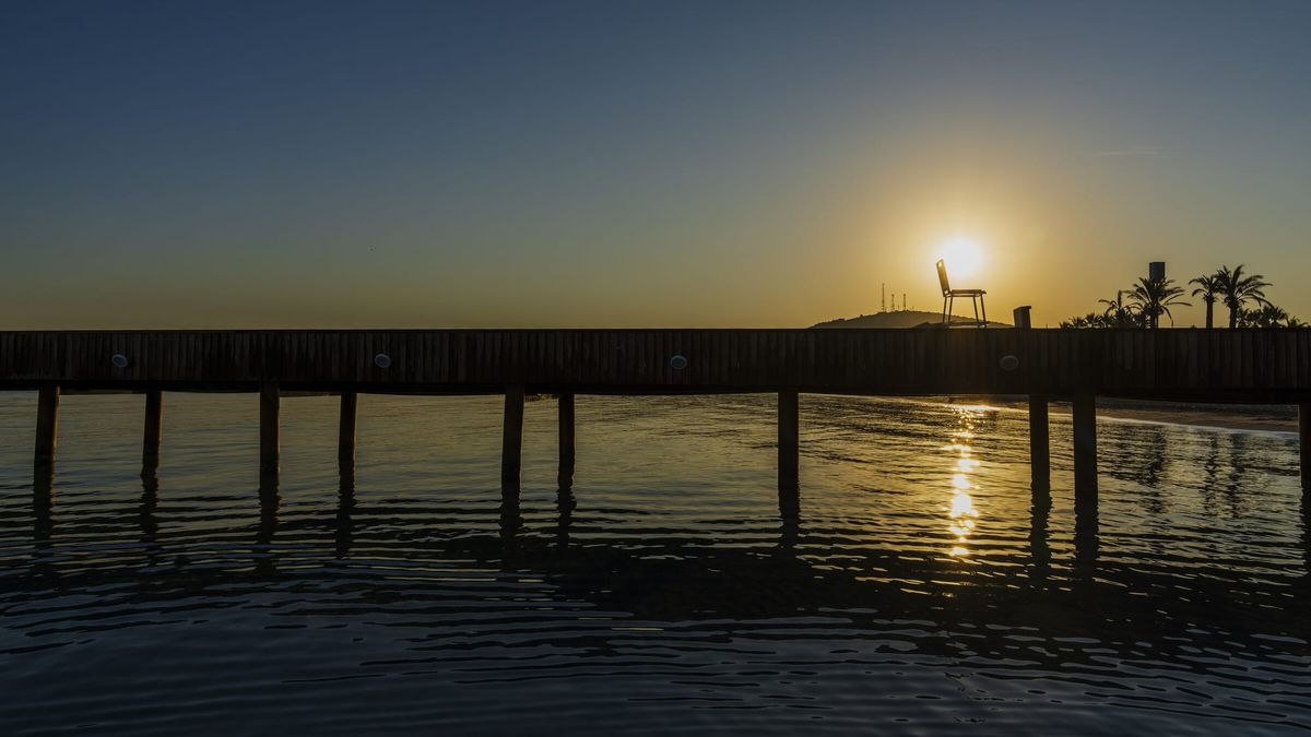 A lone empty chair silhouetted beside the setting sun at the end of a long jetty over calm, still water