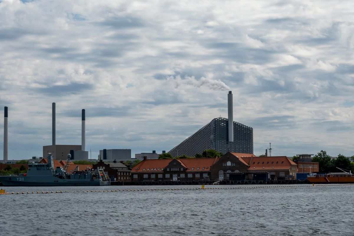 A Copenhagen waterfront with power-station chimneys and the CopenHill waste-to-energy plant releasing a plume of steam, rising behind the red-roofed buildings of an old naval yard