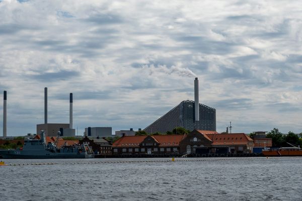A Copenhagen waterfront with power-station chimneys and the CopenHill waste-to-energy plant releasing a plume of steam, rising behind the red-roofed buildings of an old naval yard