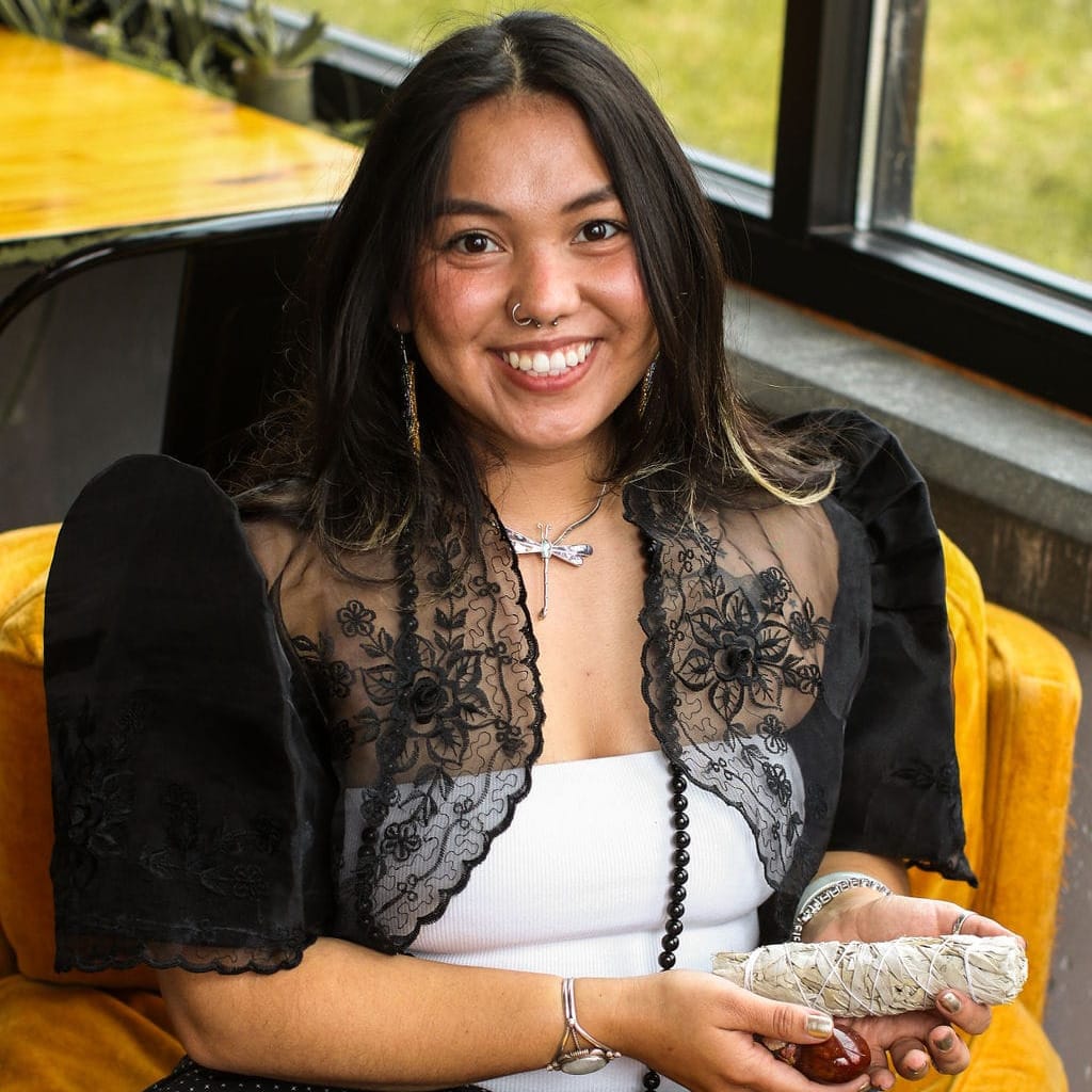 A person dressed in cultural regalia sits holding a bundle of sacred medicines including sage and minerals. 