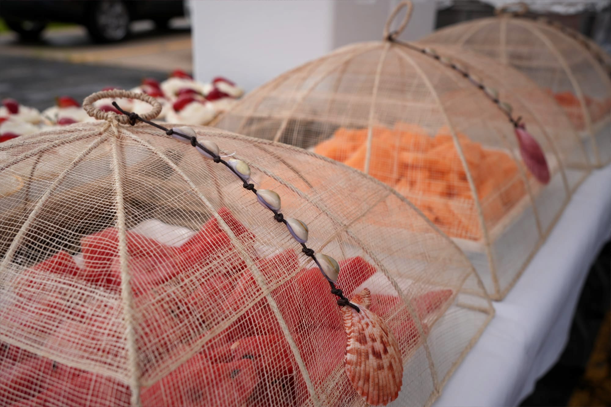 Trays of sliced fruit, including watermelon and cantaloupe, sit atop a white clothed table. On top of the trays are rattan covers with a strand of shell decorations. Behind the fruit trays are strawberry cupcakes.