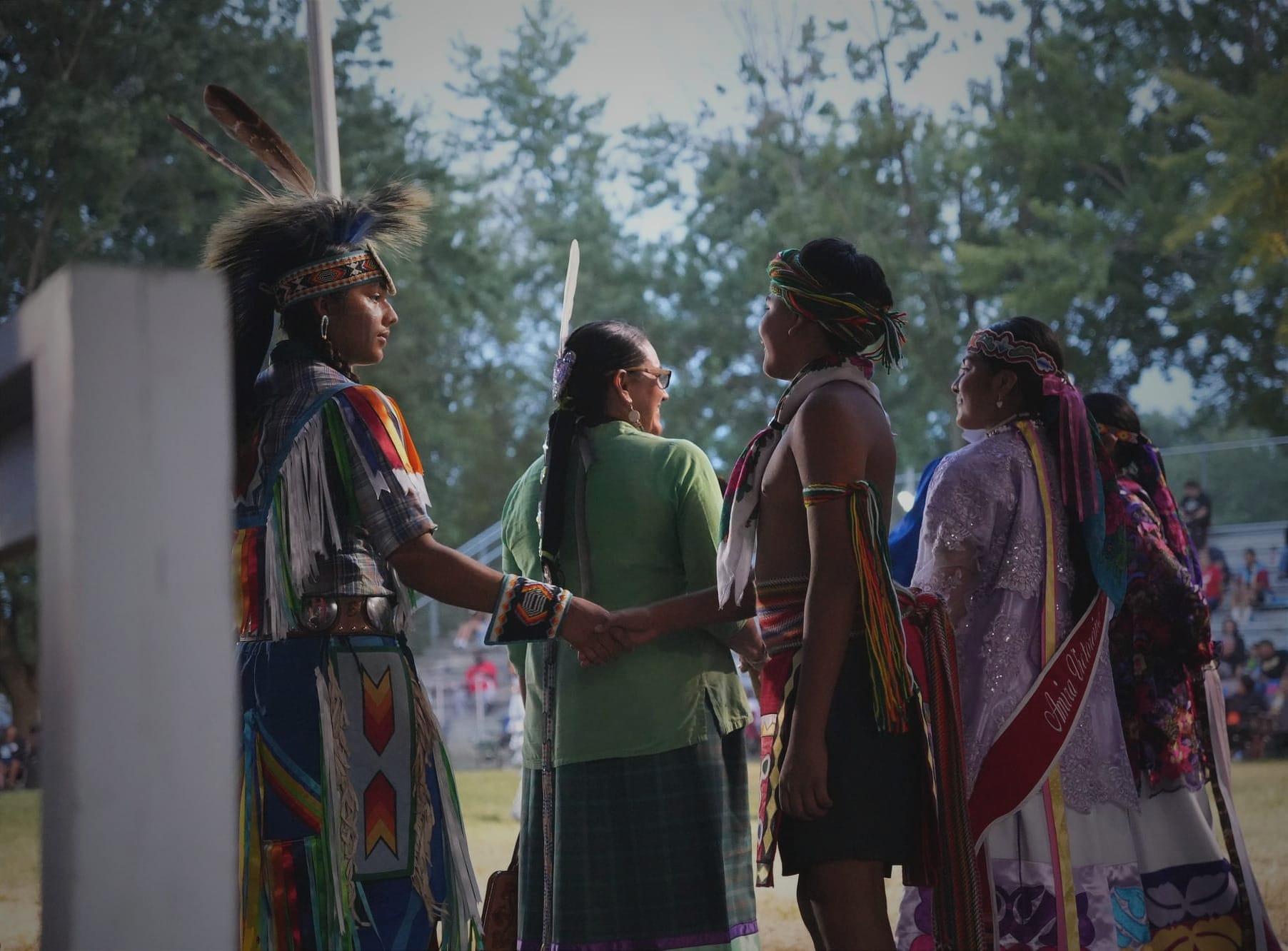 Five individuals dressed in colorful regalia are standing together before entering the powwow grounds. Two young men shake hands as three women are smiling at one another behind them. 