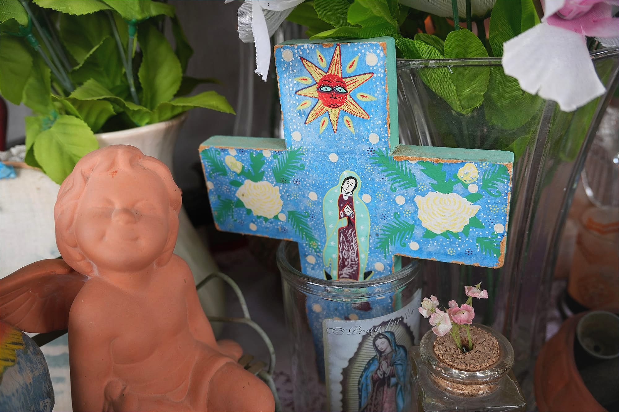 A terracotta cerub smiles looking upward with its eyes closed as it sits next to a an empty candle glass with an image of Our Lady of Guadalupe. In the candle glass is a painted blue cross with roses on the left and right sides, a yellow and orange sun with a face, and a illustration of Our Lady of Guadalupe in the middle. In the background, there appears to be vases of flowers.