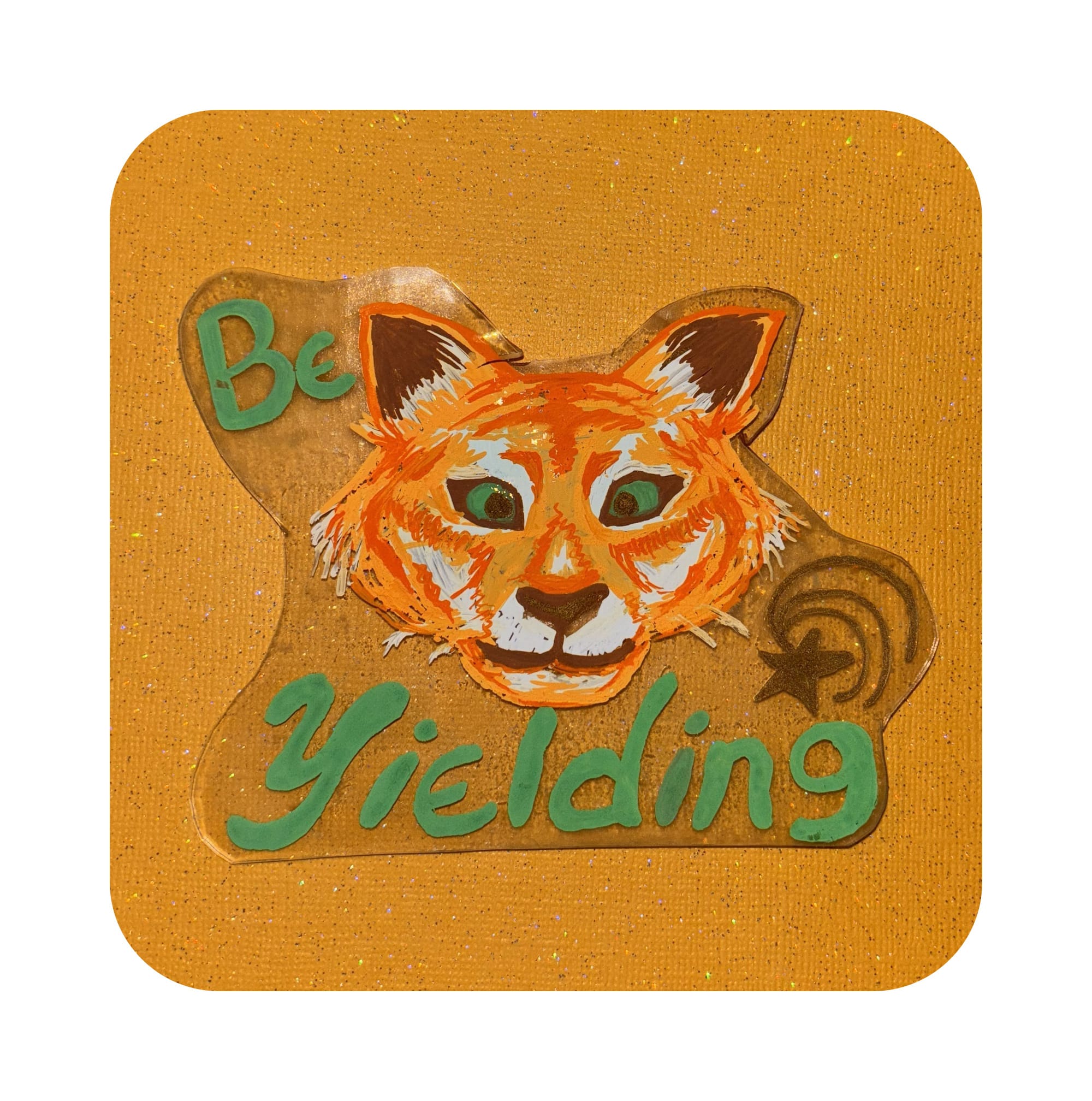 On a orange glitter background is a clear magnet painted with a white and orange tiger with brown details for the ears, nose and mouth. It has teal eyes that match the color of text that reads be yielding. Next to the tiger is a star with a swirl marks radiating out from it. 