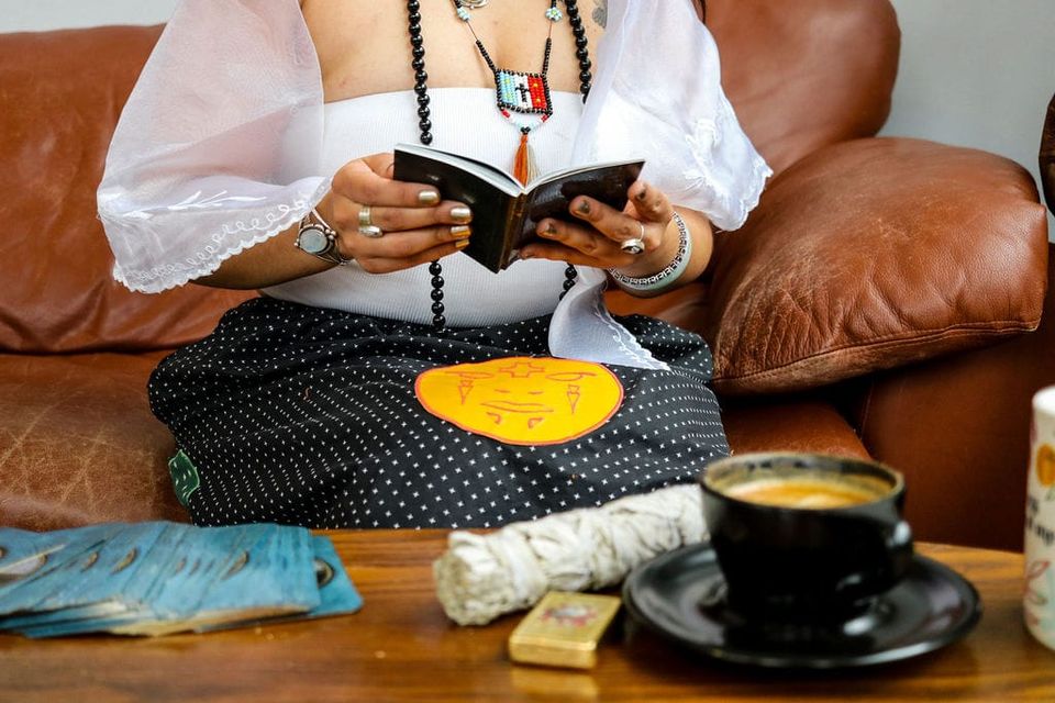 A person dressed in cultural regalia sits reading a small book in front of a coffee table that features a coffee mug, bundle of sage, a lighter, and a spread out deck of cards.