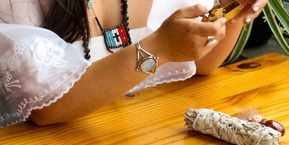 A close up of the hands of a person in cultural regalia holding a lighter while there is a bundle of sage in front on them on a table.