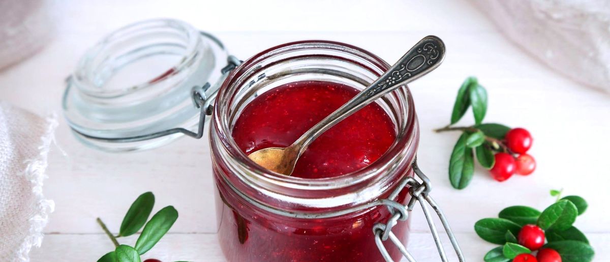 An open jar of red berry jam with a spoon in it, on a white wooden table