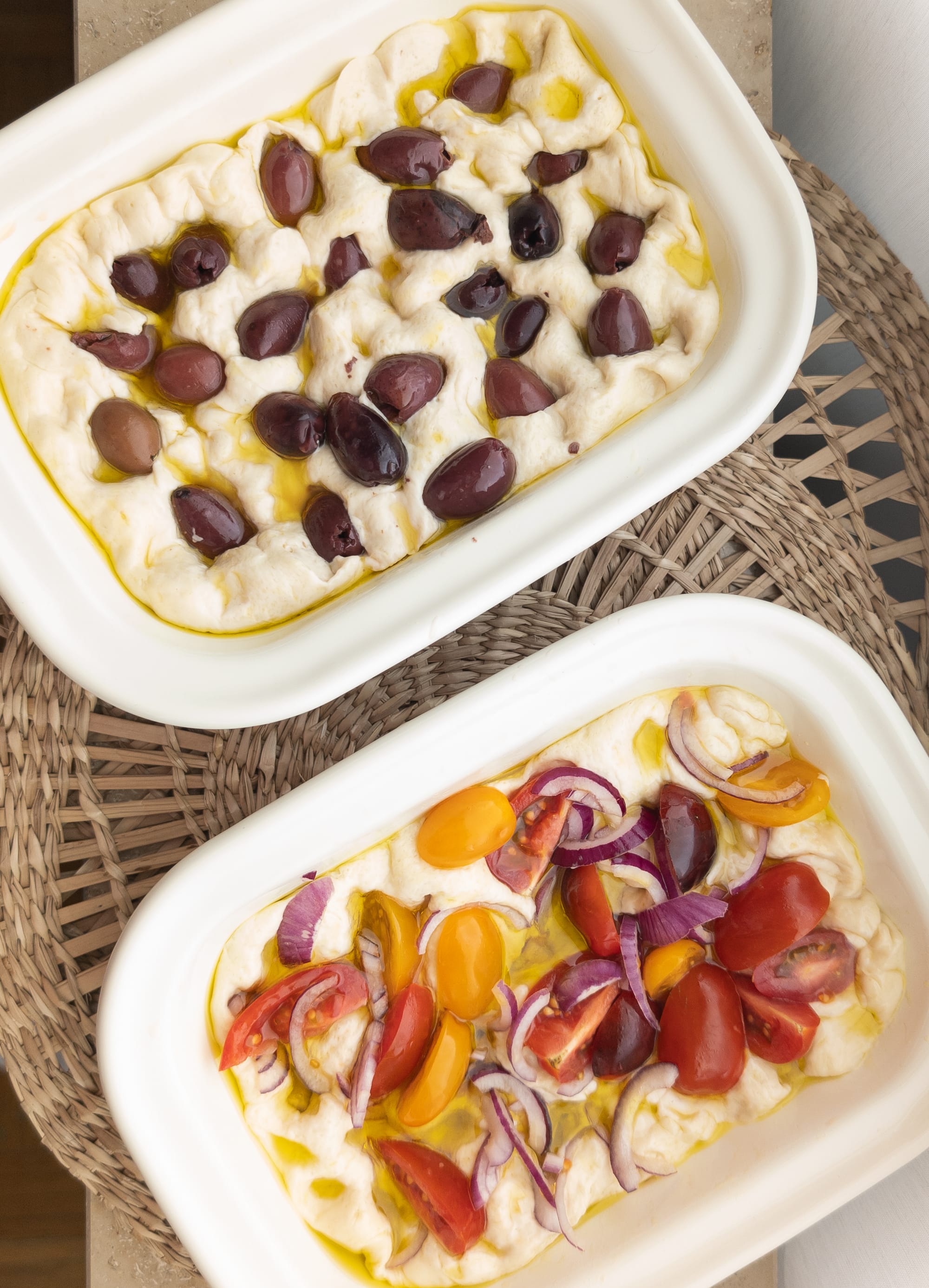 Zwei Bleche mit Focaccia. Eine belegt mit Oliven, die andere mit Cherrytomaten und roten Zwiebeln. 