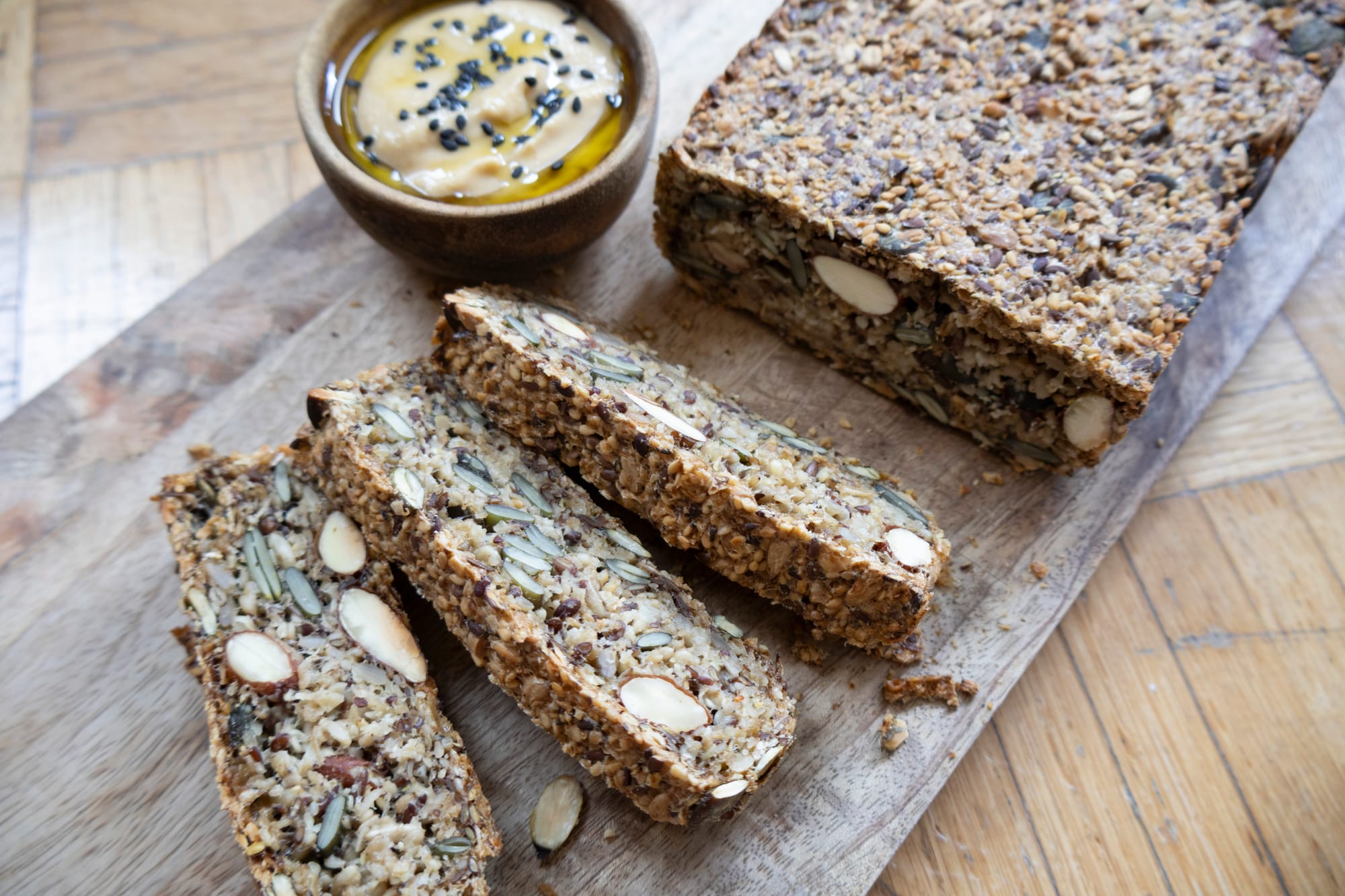 Teilweise aufgeschnittenes Saatenbrot auf Holzbrett, dazu ein Schälchen mit Hummus