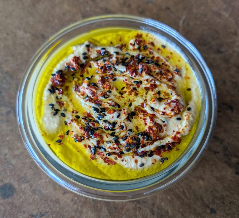 Eine Schale mit cremigem Hummus garniert mit Olivenöl, Chiliflocken und schwarzen und weißen Sesam