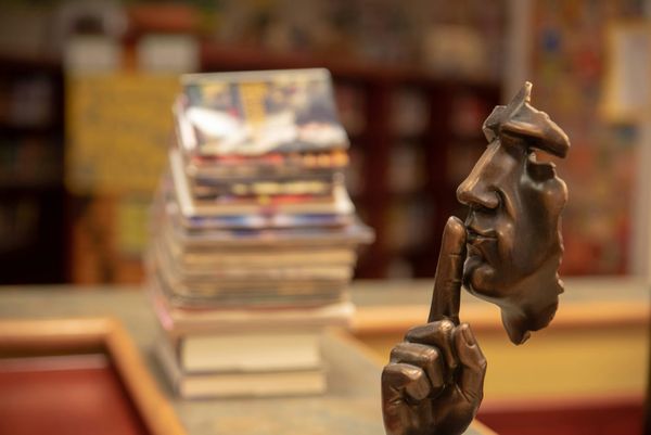 A photo of a sculpture of a man's face, in profile, holding a finger to his lips. In the background, books are out of focus