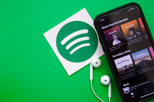 On the right, an angled smartphone showing Spotify open and showing playlists. To its left, a printed cutout of the Spotify logo on white paper and wired earbuds against a green background