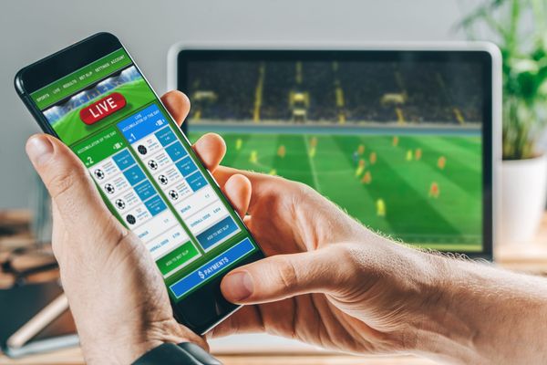 A hand holding a smartphone with a sports betting app open against the backdrop of a screen showing soccer