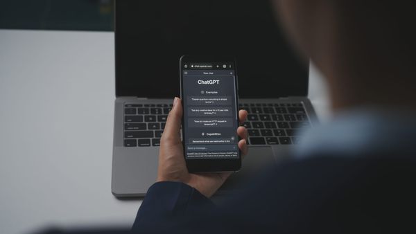 ChatGPT showing on a smartphone being held in someone's hand against the backdrop of an open MacBook.