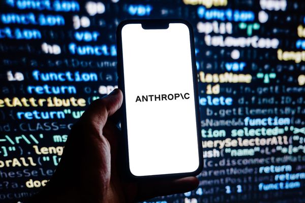 The Anthropic wordmark on a white background, shown on a smartphone in a person's hand against a backdrop of code on a monitor