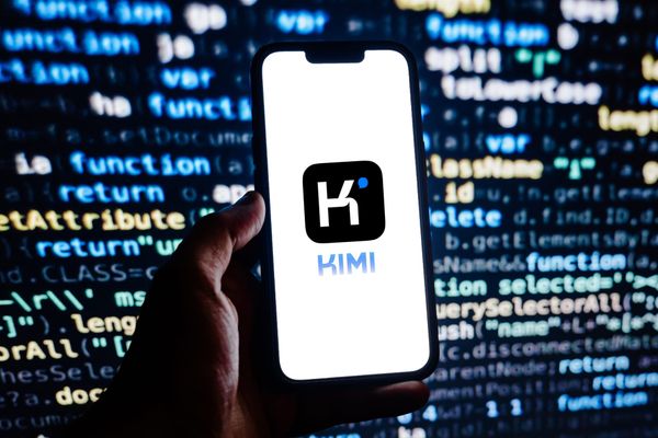 The Kimi logo and wordmark on a smartphone held in a man's hand against a backdrop of code