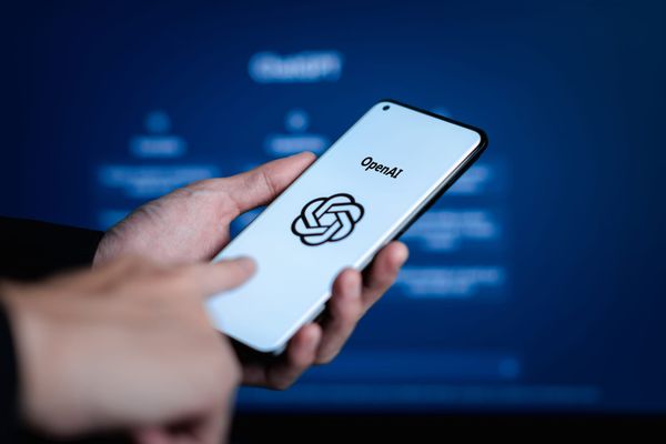 A hand holding a smartphone showing the OpenAI wordmark and logo on a white background against a blue backdrop.
