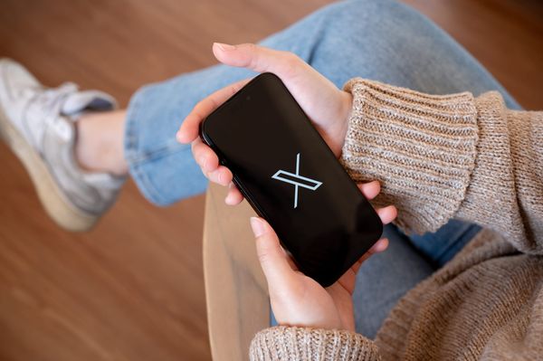 The X logo showing on a black screen on a smartphone held by a person wearing a brown sweater and blue jeans