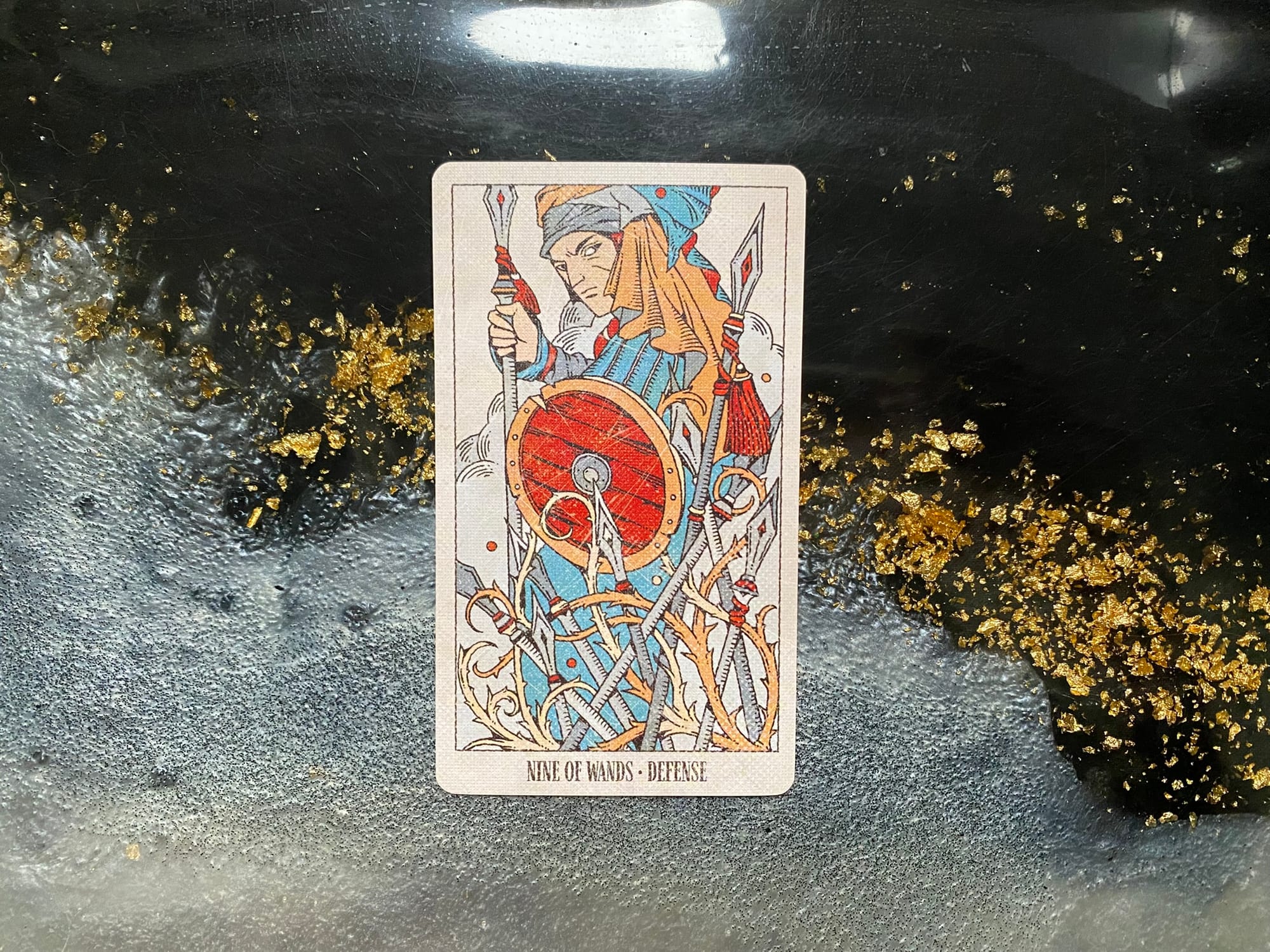 This image shows a tarot card labeled "Nine of Wands - Defense" displayed on a black surface with decorative gold leaf or gold flakes scattered around it. The card depicts a warrior figure wearing orange and blue garments with a turban-like head covering. The character is holding a staff and stands behind a red shield. Several wands or staffs appear crossed in the background, creating a defensive barrier pattern. The card has a light-colored border and the card name "NINE OF WANDS • DEFENSE" is printed at the bottom. The artistic style is from the Legendarium tarot deck with detailed line art and color accents in red, orange, and blue.