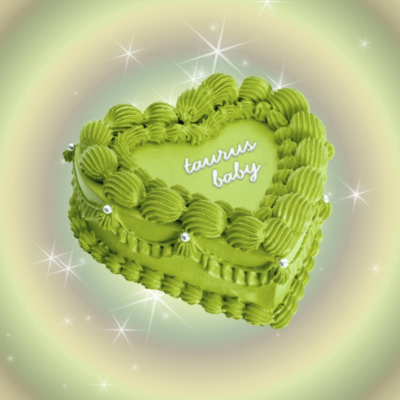 A heart-shaped cake with bright lime green frosting decorated with piped buttercream swirls around the edges. The cake has 'taurus baby' written in white script across the center and is adorned with small silver pearl decorations.
