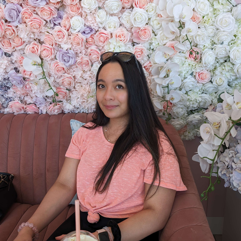 Nadine smiling softly in front of a wall of pink, white, and lavender roses, wearing a pink top, long dark hair down, sunglasses resting on her head.