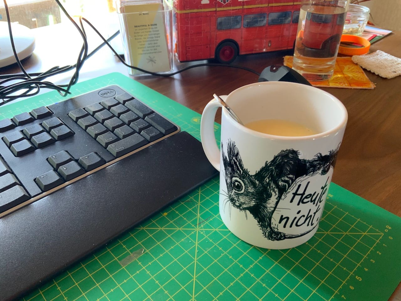 Tastatur auf grüner Schneidematte. Davor eine Tasse mit gelblichem Inhalt (Pudding). Auf der Tasse ein Eichhörnchen. Im Hintergrund Deko und ein Glas Wasser.