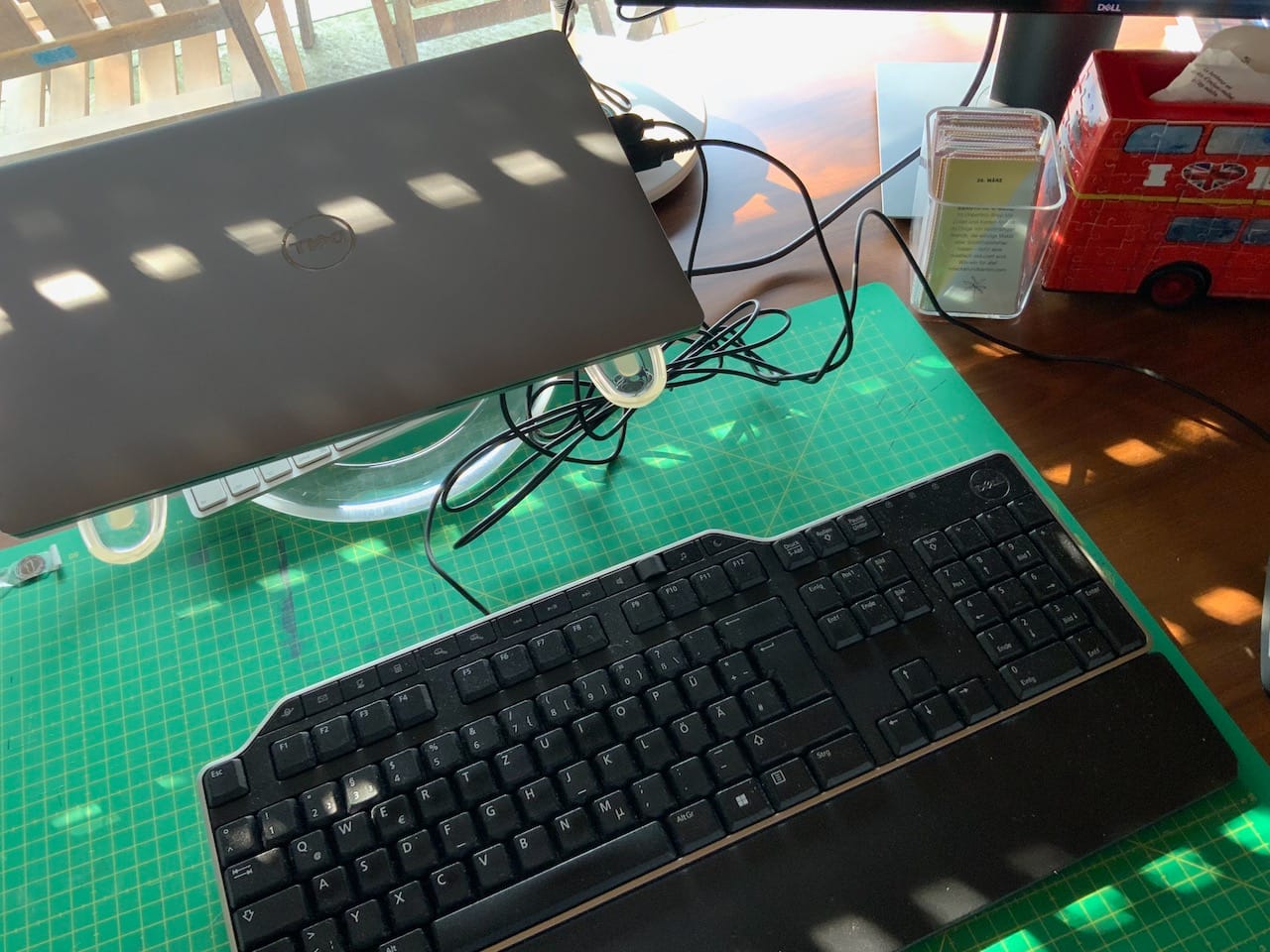 Tastatur von oben auf grüner Schneidematte. Zugeklappter Laptop von oben. Sonnenlicht fällt durch die Rollade.