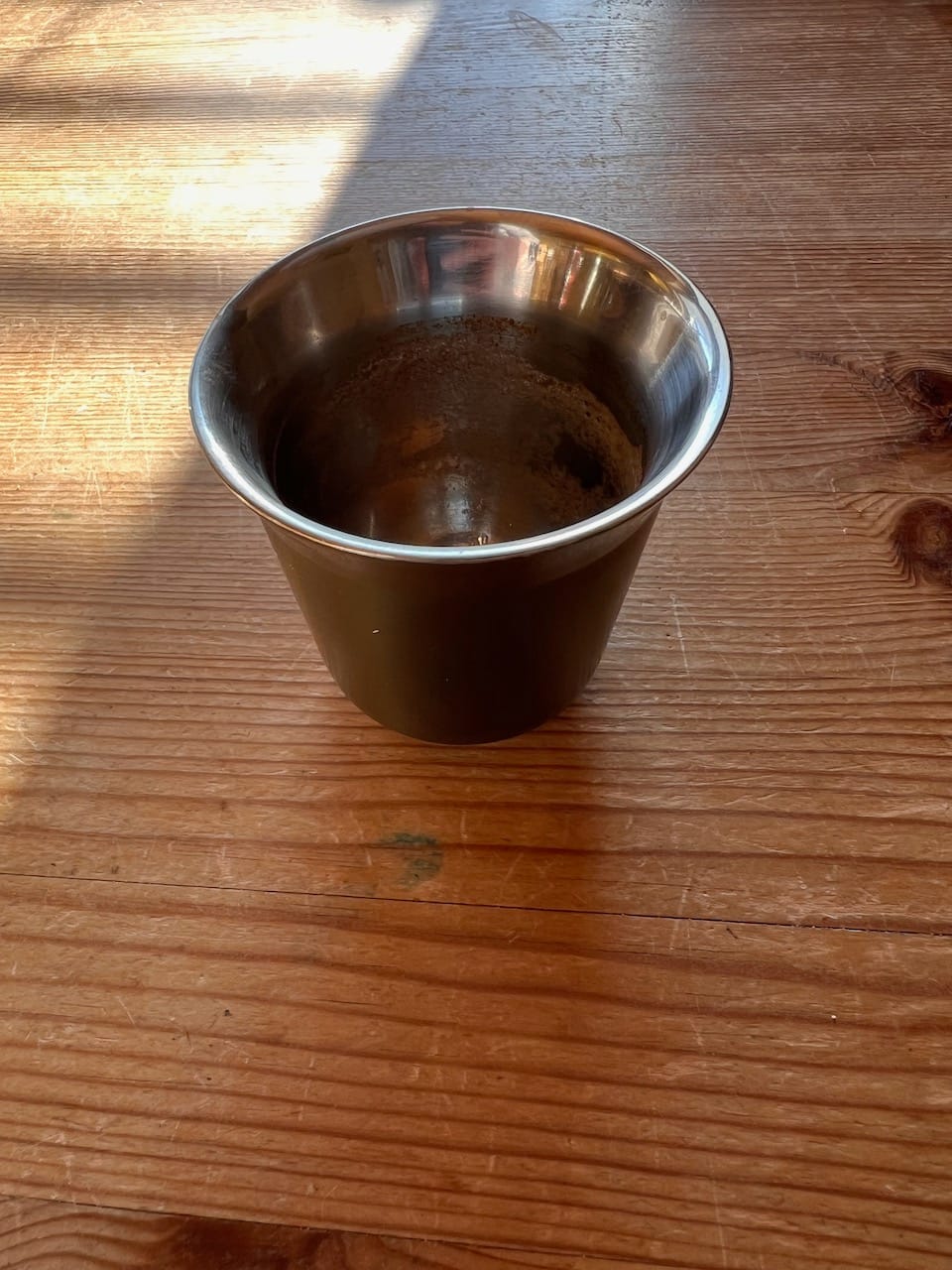 kleiner leerer Edelstahlbecher mit Kaffeeresten auf einem Holztisch. Sanftes Licht fällt auf die Tischplatte.