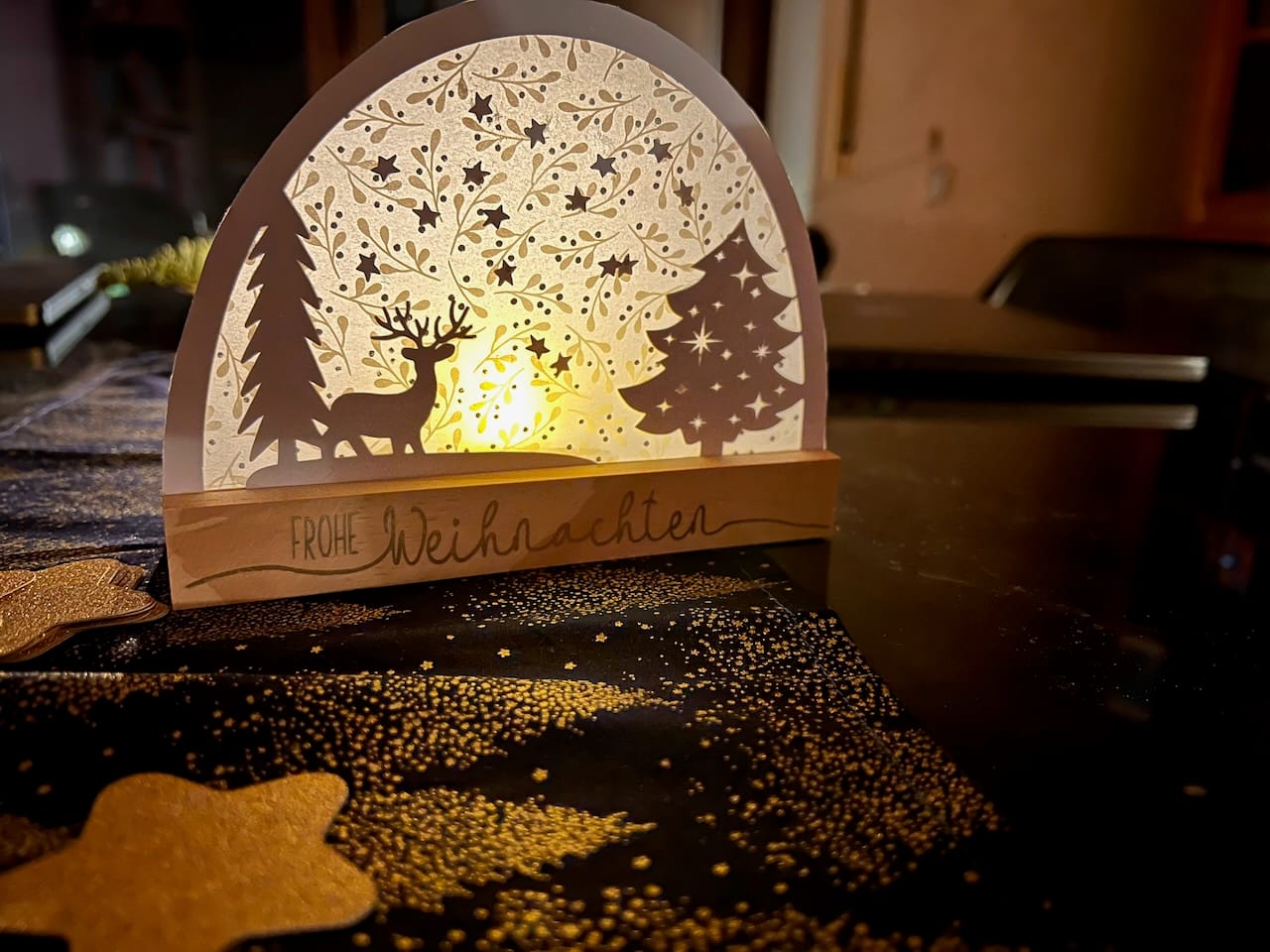 Bogen aus Papier mit Tannen und Hirsch, Transparentpapier und einem LED-Teelicht dahinter. Der Bogen ist in einem Holzklotz, auf dem Frohe Weihnachten steht.