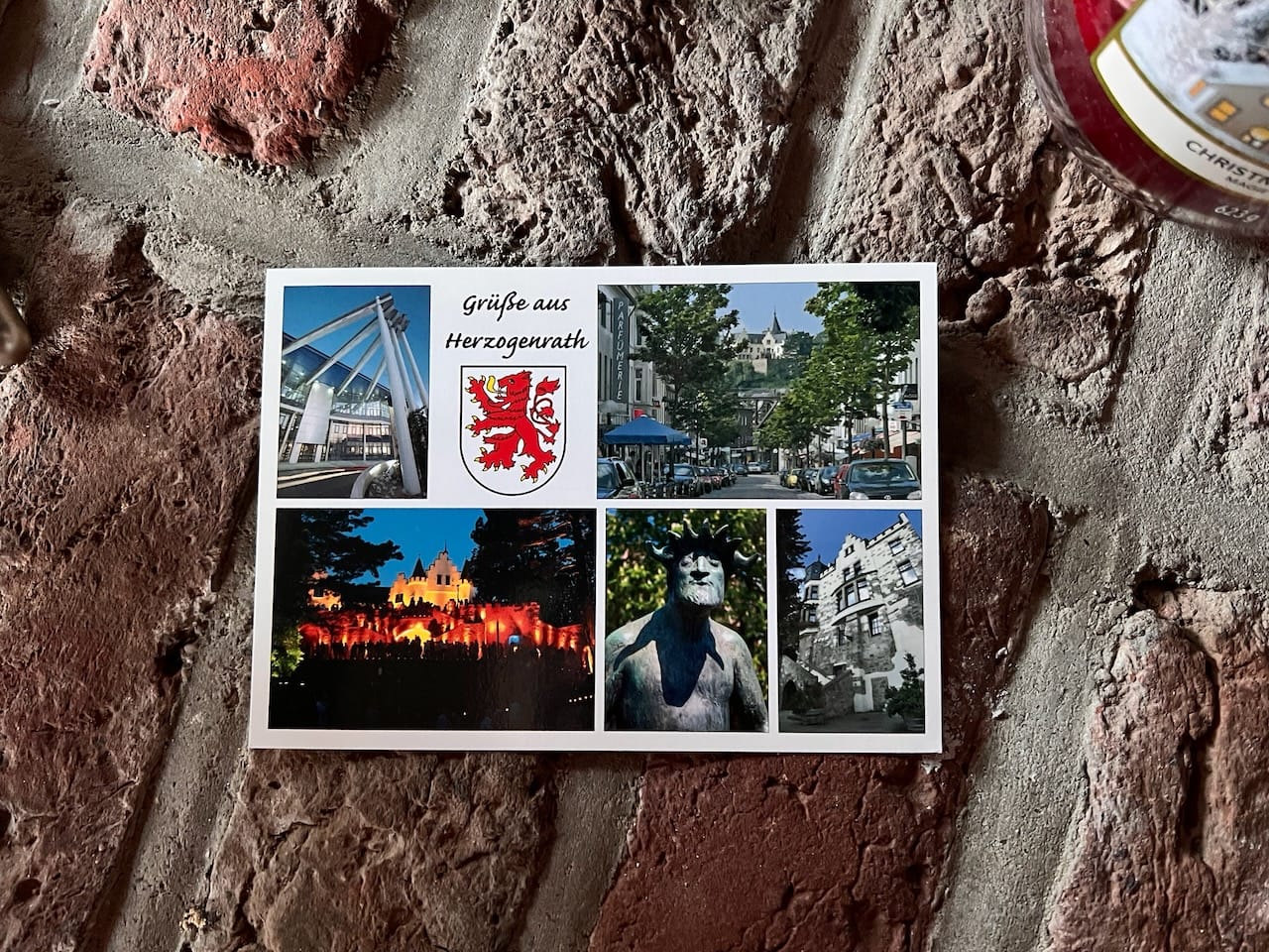 Postkarte mit fünf Bildern und dem Schriftzug „Grüße aus Herzogenrath“ mit dem Stadtwappen. Die Bilder haben alle einen Bezug zur Stadt Herzogenrath (z. B. eine Statue oder die bunt angestrahlte Burg Rode). 