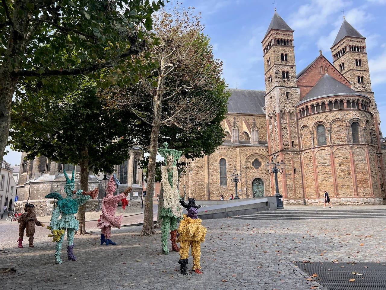 Eine Kirche im Hintergrund auf einem Platz. Bäume an der linken Seite, neben den Bäumen bunte, teilweise hohe, Skulpturen