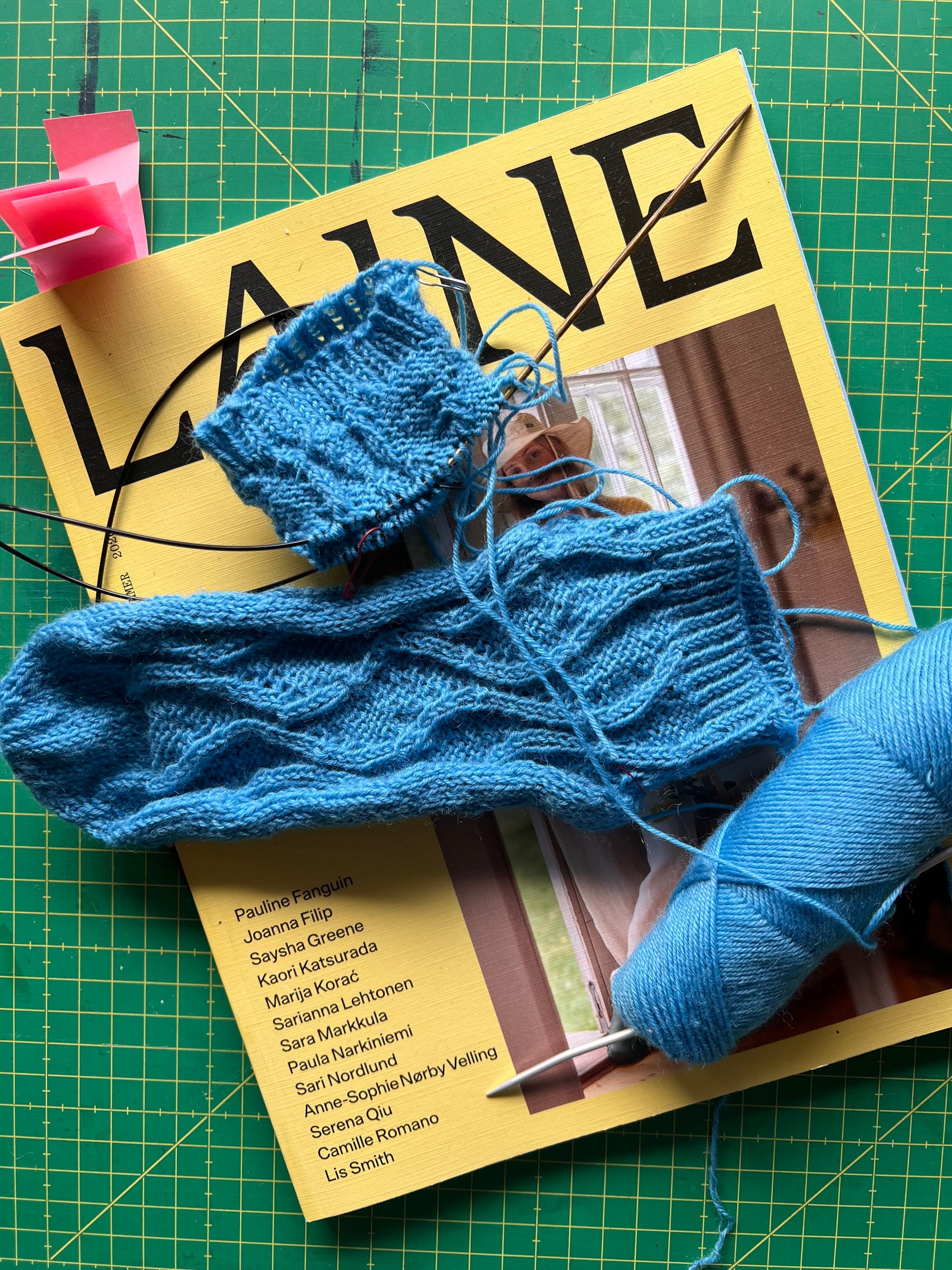 ein fertiger Sock in blau mit Wellenmuster und ein begonnener Sock liegen gemeinsam mit dem Knäuel Wolle auf einer gelben Zeitschrift. Die Zeitschrift hat den Titel "Laine"