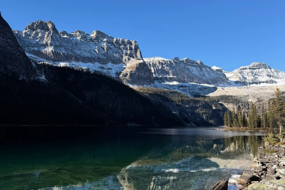 Clear green water beside Boom Lake's rocky shore, with evergreen forest and snow-dusted cliffs reflected in the lake.