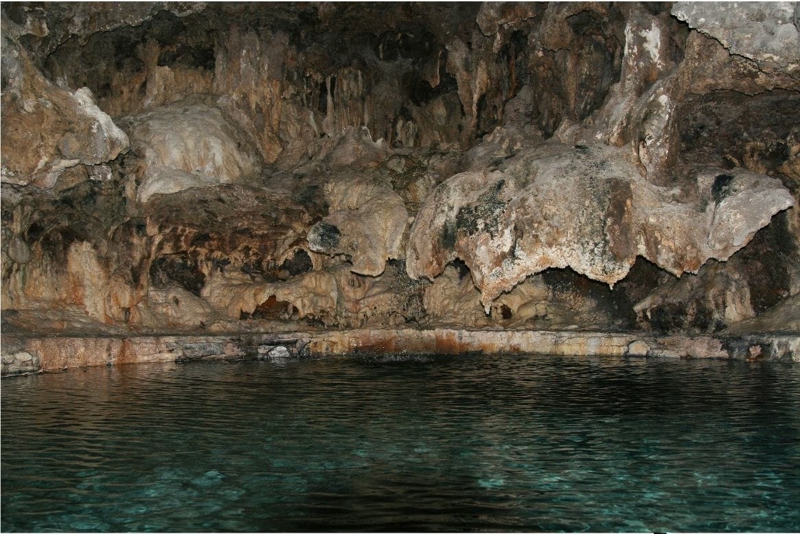 Grey and brown rock formations overhang a dark green pool inside the cave at Cave and Basin National Historic Site.
