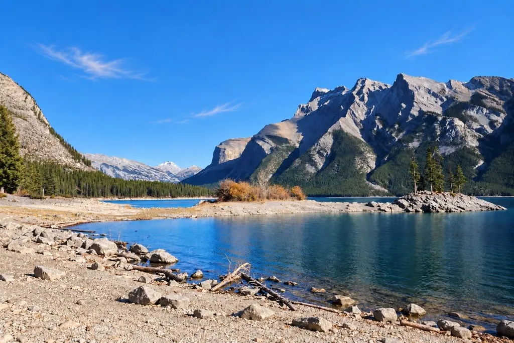 Rocky Lake Minnewanka shoreline with driftwood, blue water and steep mountain slopes beyond.