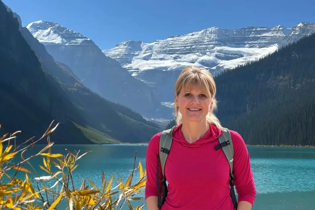 A woman wearing a pink top and a backpack beside Lake Louise, with snow-covered mountains behind her.