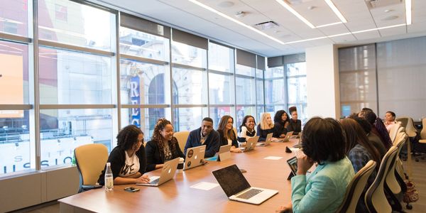 about twenty people, all with laptops or tablets, meeting at a large table in a room with a large window