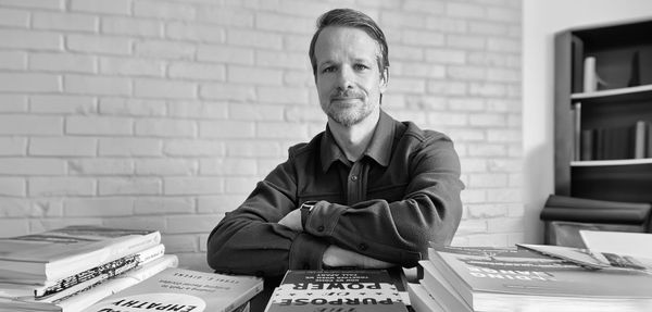 a black-and-white photo of a man with a light beard sitting at a table, facing the viewer, surrounded by stacks of books